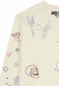 Chemise crème à manches longues avec un dessin au trait coloré incluant deux colibris, un chérubin, un crâne aux yeux en forme de cœur, un flamant rose, un soleil, un signe de paix et des fleurs.