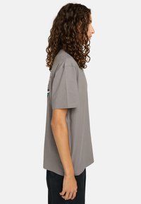 Joven con cabello rizado que lleva una camiseta gris holgada y pantalones negros, de pie de perfil contra un fondo blanco liso.