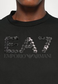 Melnas kokvilnas t-krekls ar sequīnu "EA7" logotipu un "EMPORIO ARMANI" tekstu eleganta dizainā, ar gludu tekstūru un apaļu kakla izgriezumu.