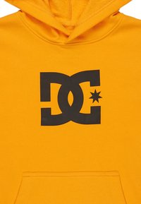 Žltá mikina z mäkkej látky s veľkým čiernym logom DC a grafickým motívom hviezdy na prednej strane. Obsahuje klokanie vrecko.