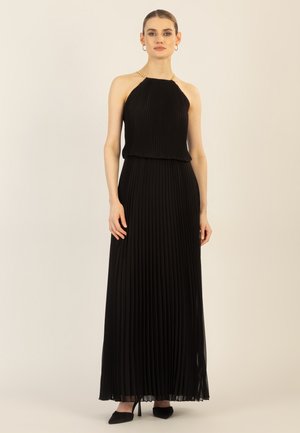 Frau steht in einem ärmellosen, plissierten schwarzen Neckholder-Kleid mit goldenen Kettenriemen und schwarzen spitzen Absätzen vor einem schlichten Hintergrund.