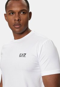 T-shirt bianca in cotone con scollo rotondo e maniche corte, con il logo nero EA7 Emporio Armani sul lato sinistro del petto.