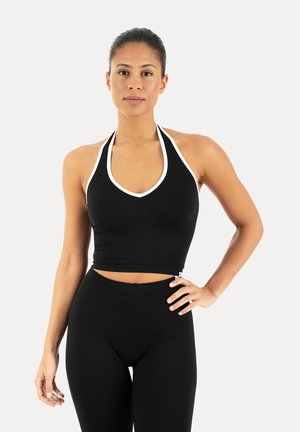 Kobieta z zaczesanymi do tyłu włosami, ubrana w czarny bezrękawnik halter z białym wykończeniem oraz czarne legginsy z wysokim stanem, stojąca z ręką na biodrze.