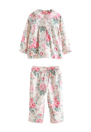 Junges Mädchen lächelt und steht vor einer blumigen Tapete, trägt passende rosa-weiße geblümte Pyjamas.