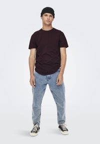 Burgundy short-sleeve T-shirt, classic fit, worn with light blue, loose-fitting jeans and black sneakers. Model wearing a black beanie.