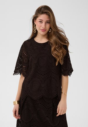 Femme aux longs cheveux ondulés porte un haut en dentelle noire et une jupe avec des motifs circulaires, des boucles d’oreilles créoles dorées ainsi qu’un bracelet et des bracelets assortis.