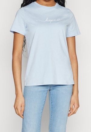 Femme portant un t-shirt bleu clair avec le mot "hope" et un petit cœur au-dessus d'un jean bleu clair.