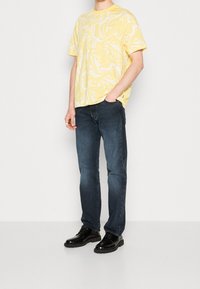 Person som bär en kortärmad skjorta i gult och vitt med virvlar, mörkblå jeans, svarta läderskor, stående med en hand i fickan.