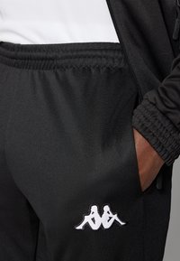 Black athletic pants with elastic waist and cuffs, featuring a white embroidered logo on the pocket. Made of smooth, stretchy fabric.
