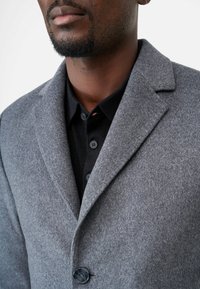 Grijze wolmix jas met een gestructureerde kraag, een enkele knoopsluiting en een soepele textuur, gestapeld over een zwart polohemd met knopen.