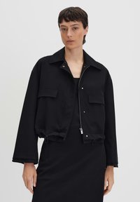 Donna con capelli corti e scuri che indossa una giacca nera cropped con tasche frontali e zip, in piedi davanti a uno sfondo chiaro uniforme.