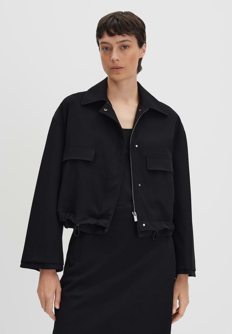 Donna con capelli corti e scuri che indossa una giacca nera cropped con tasche frontali e zip, in piedi davanti a uno sfondo chiaro uniforme.