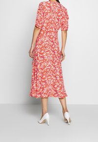 Robe midi rouge à fleurs avec des manches courtes bouffantes, taille cintrée et jupe fluide. Portée avec des chaussures à talons hauts blanches. Tissu texturé.