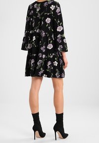 Czarna sukienka w kwiaty z długimi dzwonowymi rękawami, zdobiona białymi i lawendowymi kwiatami. Warstwowy fason z guzikiem z tyłu. Noszona z czarnymi szpilkami.