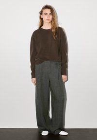 Pull en maille brun avec manches longues, associé à un pantalon gris ample. Le modèle porte des baskets blanches. Design simple et minimaliste.