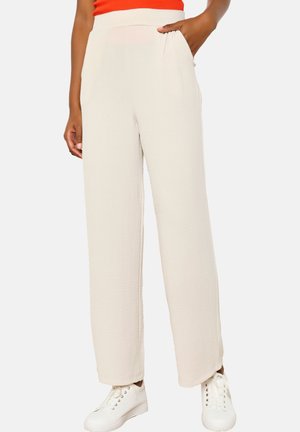 Hoge taille, wijde broek in een lichtbeige stof met een textuurafwerking. Heeft zijdelingse zakken en subtiele plooien. Gecombineerd met witte sneakers.