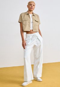 Zkrácená béžová košile s krátkými rukávy, bílý top s úzkými ramínky a bílé široké kalhoty. Světlé tenisky dotvářejí outfit. Jednoduché pozadí.