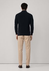 Man i svart långärmad tröja, beige byxor och bruna skor som står vänd mot en ljusgrå vägg, med händerna avslappnat vid sidorna.