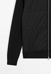 Giacca nera con pannello frontale trapuntato e maniche in maglia. Include una cerniera argentata e tasche laterali. Texture liscia con un design casual.