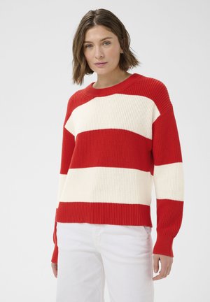 Nő, piros-fehér csíkos kötött pulóvert és fehér nadrágot visel, egyszerű világos háttér előtt áll.