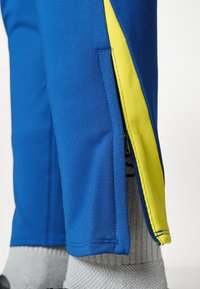 Μπλε αθλητικό παντελόνι με κίτρινη ρίγα στο πλάι. Διαθέτει φερμουάρ κοντά στο στρίφωμα και μαλακό, υφασμάτινο υλικό. Γκρι κάλτσες ορατές πάνω από τον αστράγαλο.