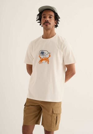 Mann trägt beige Mütze, weißes T-Shirt mit orangefarbener Krabben- und Schiffsgrafik und braune Cargo-Shorts, steht mit verschränkten Händen am Rücken.