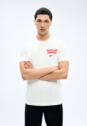 Homme aux cheveux courts et bouclés, les bras croisés, portant un t-shirt blanc avec un texte rouge "HAPPY AND GLORIOUS" et un logo Nike rouge.