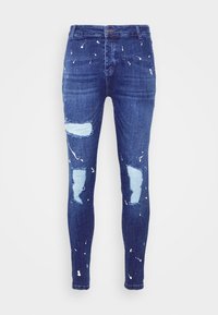 Pantalones vaqueros de mezclilla azul oscuro con roturas desgastadas, parches en azul claro y salpicaduras de pintura blanca. Ajuste slim con cierre estándar de botón y cremallera.