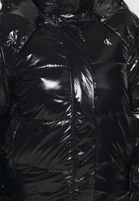 Chaqueta acolchada negra con acabado brillante, cierre de cremallera, capucha sobredimensionada y logo CK en el pecho. Diseño acolchado con costuras.