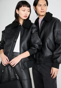 Μαύρες δερμάτινες μπόμπερ ζακέτες με χνουδωτούς μαύρους γιακάδες, ριμπ μανίκια και τσέπες με φερμουάρ, φορεμένες πάνω από λευκά μπλουζάκια και σκούρα παντελόνια.