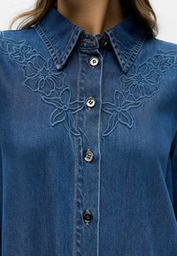 Gros plan sur une chemise en denim avec broderie florale près du col et boutons noirs sur le devant.