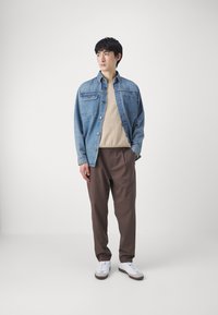 Veste en denim, bleu clair, avec deux poches poitrine ; pull beige en dessous ; pantalon plissé marron ; baskets blanches avec des accents gris.