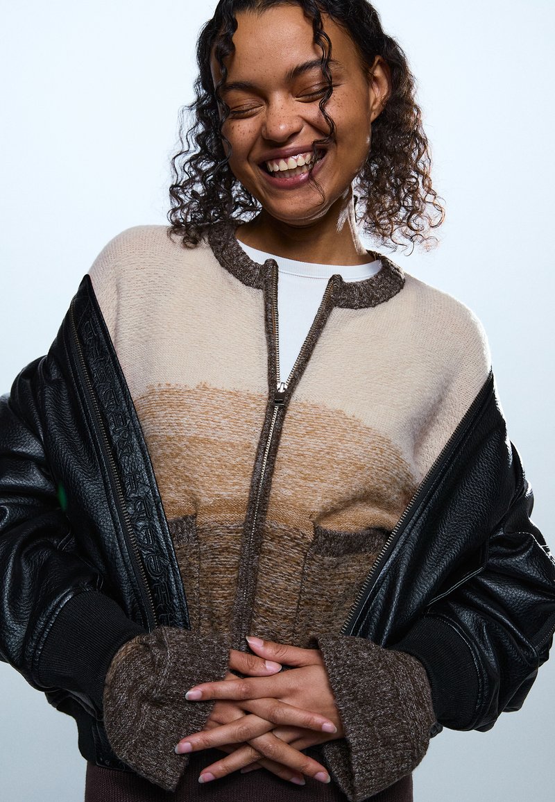 Lächelnde Frau mit lockigem Haar trägt einen beigen und braunen Reißverschlusspullover unter einer schwarzen Lederjacke, die Hände vor der Taille verschränkt, die Augen geschlossen vor einem schlichten Hintergrund.