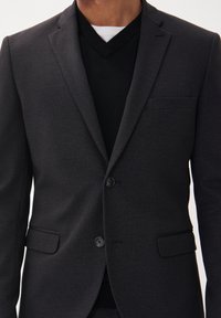Tumehall blazer, mis on valmistatud tekstureeritud kangast, kahe nupuga, ühe rinnataskuga ja terava kraega. Kantud musta V-kaelusega särgi peal.