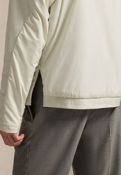 Αθλητικό μπ ουφάν ανοιχτού μπεζ με λεία υφή και ελαστική μέση με λογότυπο, συνδυασμένο με σκούρο γκρι παντελόνια καλοραμμένα.