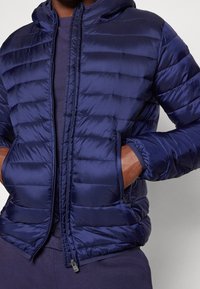 Marine blauwe pufferjack met een capuchon, voorzien van een geborduurd ontwerp, een ritssluiting aan de voorkant en zakken aan de zijkanten. Gemaakt van glanzend, lichtgewicht materiaal.