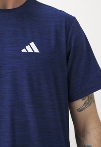 Navy blue athletic t-shirt in a textured, moisture-wicking fabric, featuring a large white logo on the left chest. Short sleeves.