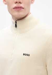 Light beige zip-up sweater with high collar and textured sleeves, featuring a black "BOSS" logo on the left chest. Soft knit material.