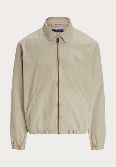 Beige Reißverschlussjacke aus einem weichen Stoff. Sie verfügt über einen Kragen, elastische Bündchen, Seitentaschen und ein kleines Logo auf der Brust.