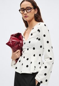 Hvit langermet topp med svarte polkadotter, V-utringning, figursydd overdel og peplumkant. Modellen holder en rød skinnclutch.