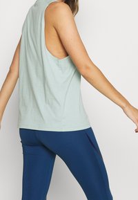 Top verde chiaro senza maniche con design a schiena aperta, realizzato in tessuto morbido. Abbinato a leggings blu navy con dettagli laterali.