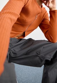 Top de manga larga naranja acanalado con cremallera y detalle recortado, combinado con pantalones cargo gris oscuro. Tela texturizada con costuras visibles.