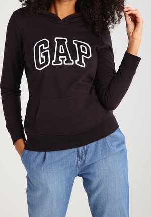 Woman wearing black GAP hoodie with white logo and light blue jeans, posing with one hand in pocket and the other touching hair.