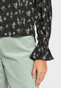 Blusa preta com flores e bolinhas brancas, com punhos franzidos. Combinada com calças de veludo cotelê verde claro texturizadas. Padrões subtis em ambas.