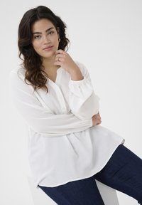 Weiße strukturierte Bluse mit lockerer Passform, Knopfverschluss und elastischen Bündchen, kombiniert mit dunkelblauen Jeans. Einfaches und vielseitiges Design.