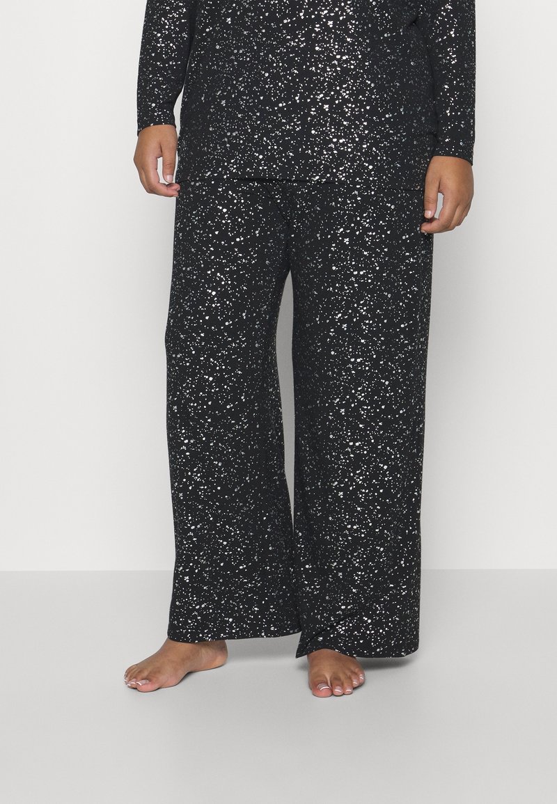 Pantalon large noir à motifs blancs tachetés, porté pieds nus, visible de la taille aux pieds sur un fond uni.