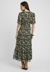 Robe maxi en tulle, motif floral vert et blanc, avec des manches courtes et un détail dos décolleté. S'associe avec des bottes marron jusqu'au genou.