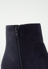 Bottine en daim bleu marine avec une finition lisse et texturée. Comprend une fermeture éclair latérale apparente et des détails cousus au niveau de la couture.