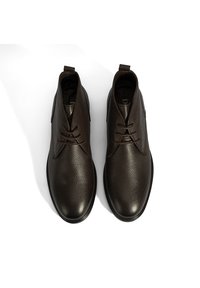 Chaussures de cheville en cuir marron foncé avec une finition texturée, présentant un orteil rond, un laçage central et des languettes au talon pour un enfilage facile.