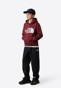 Vínová mikina s kapucňou s bielym logom, čierne tepláky a čierne tenisky. Očarujúci v neformálnej stojacej póze, nosí modrú čiapku.